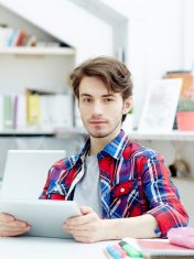 Adolescent man with touchpad sitting by desk in design studio or classroom